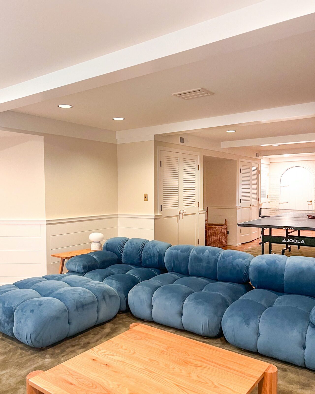 A basement family room with white and cream walls, plush green rugs, a blue modular sofa, a white oak coffee table, and a ping pong table