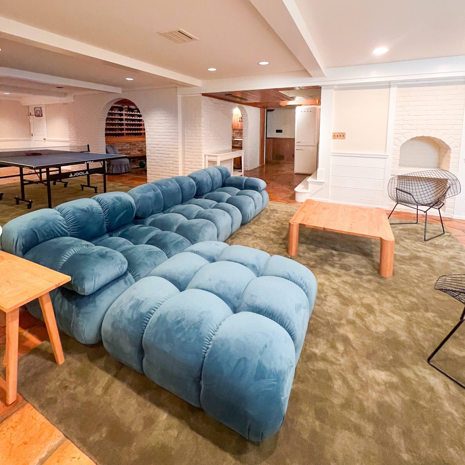 A basement family room with white and cream walls, plush green rugs, a blue modular sofa, a white oak coffee table, a ping pong table, and vintage Bertoia diamond chairs