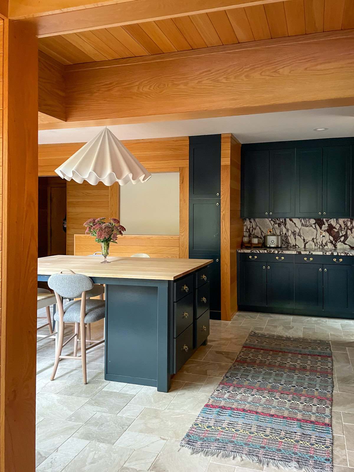 Kitchen dining area with marble flooring, an island with a butcher block countertop and a white felt pendant light over it, blue cabinets, and wood-paneled walls.