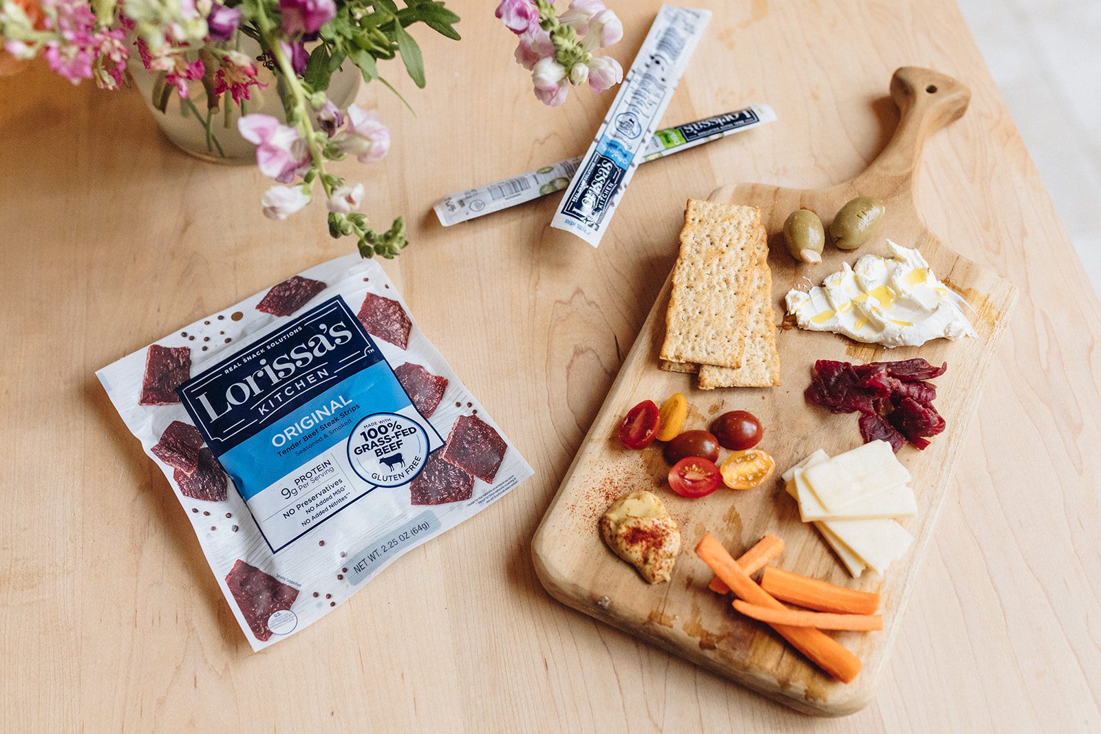 A cutting board on a butcher board with Lorissa's Kitchen packaging, along with cheese, crackers, tomatoes