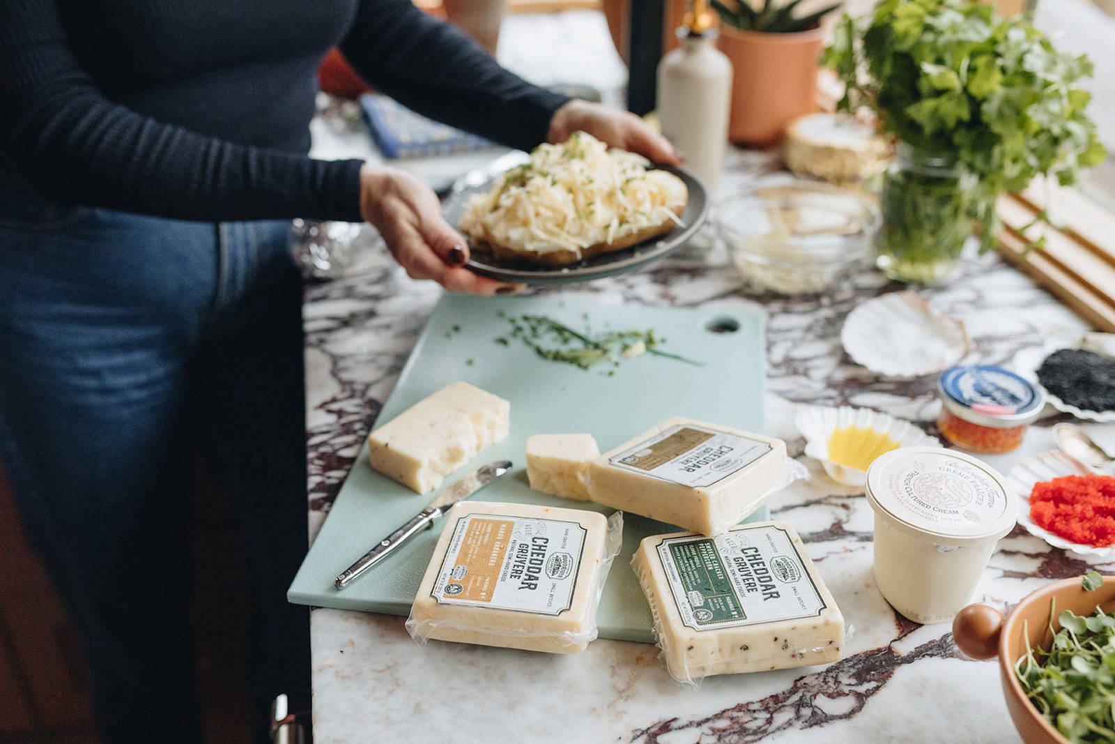 Woman preparing a meal in the kitchen with a variety of cheeses in the forefront.
