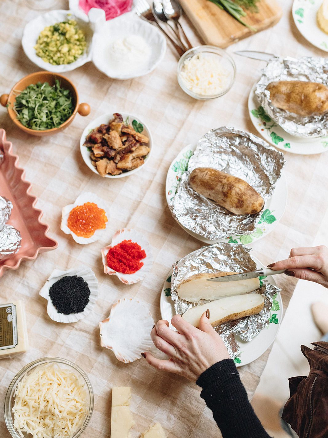 A woman slicing a baked potato in half. On the table next to her are a spread of various ingredients: cheese, cilantro, corn